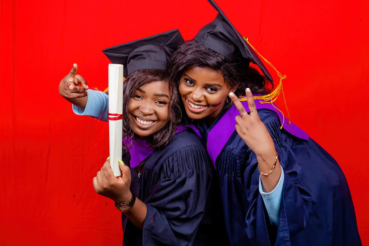 Two graduates in caps and gowns smiling with a diploma in front of a red backdrop